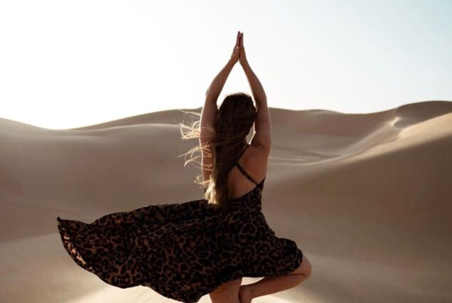 A woman in a flowing dress practicing yoga in the desert, surrounded by rolling sand dunes