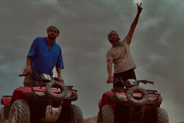 Two men on quad bikes posing in a desert under a cloudy sky