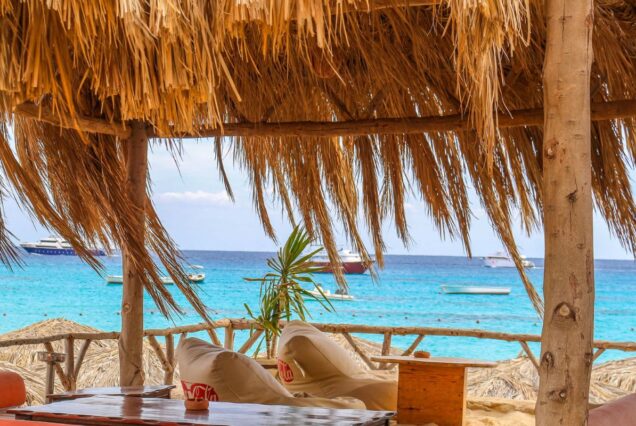 A thatched-roof seating area with a view of the turquoise sea and anchored boats