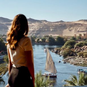 Woman admiring the scenic river view with a sailboat passing by, surrounded by desert hills