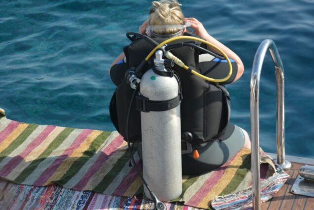 A scuba diver preparing for a dive while seated on a colorful striped mat near deep blue waters