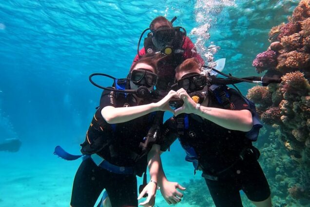 Three scuba divers underwater forming a heart shape with their hands near vibrant coral reefs